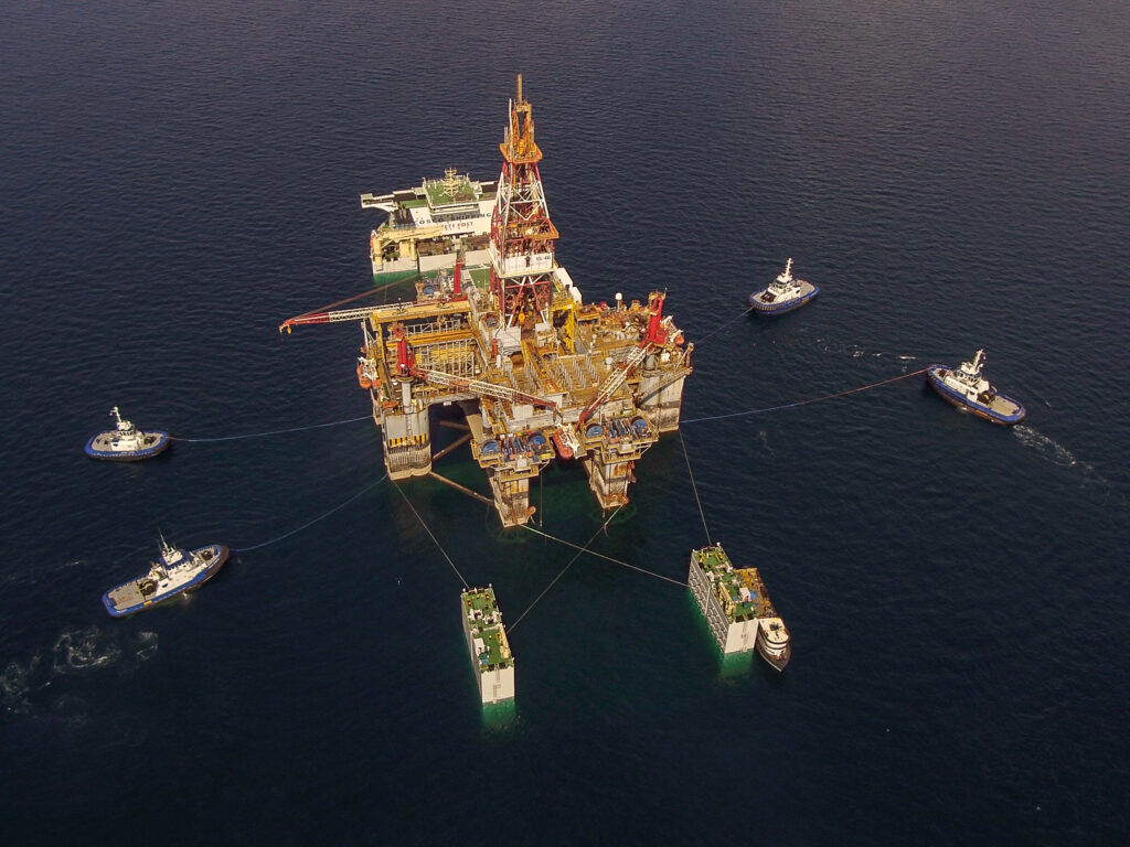 A large offshore oil rig surrounded by support boats at sea during twilight.